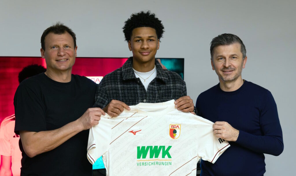 Neuzugang Kevin Nana Motowilczuk steht zusammen mit Verantwortlichen des FC Augsburg für ein Foto bereit.